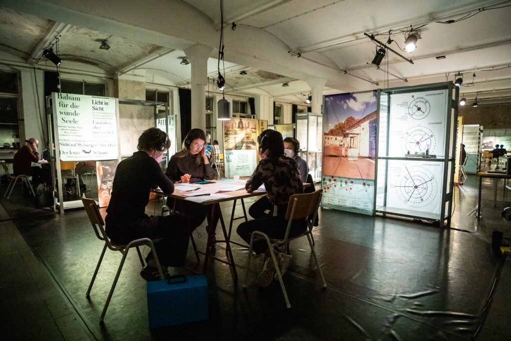 A room with several tables separated by shelves with printed material. Four people with headphones are sitting around the table in the center, which is covered with game paraphernalia.