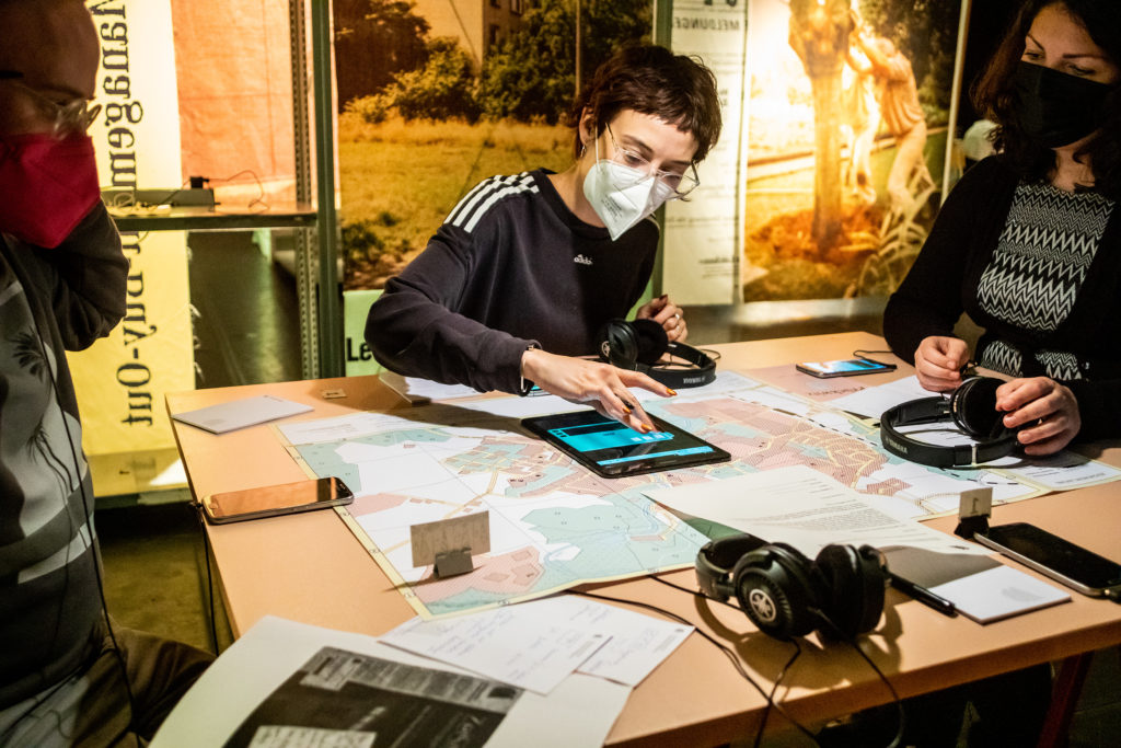 On a table are a city map, smartphones, a tablet, printed sheets of paper, and headphones. Three people are sitting around this table, one of them is using the tablet.