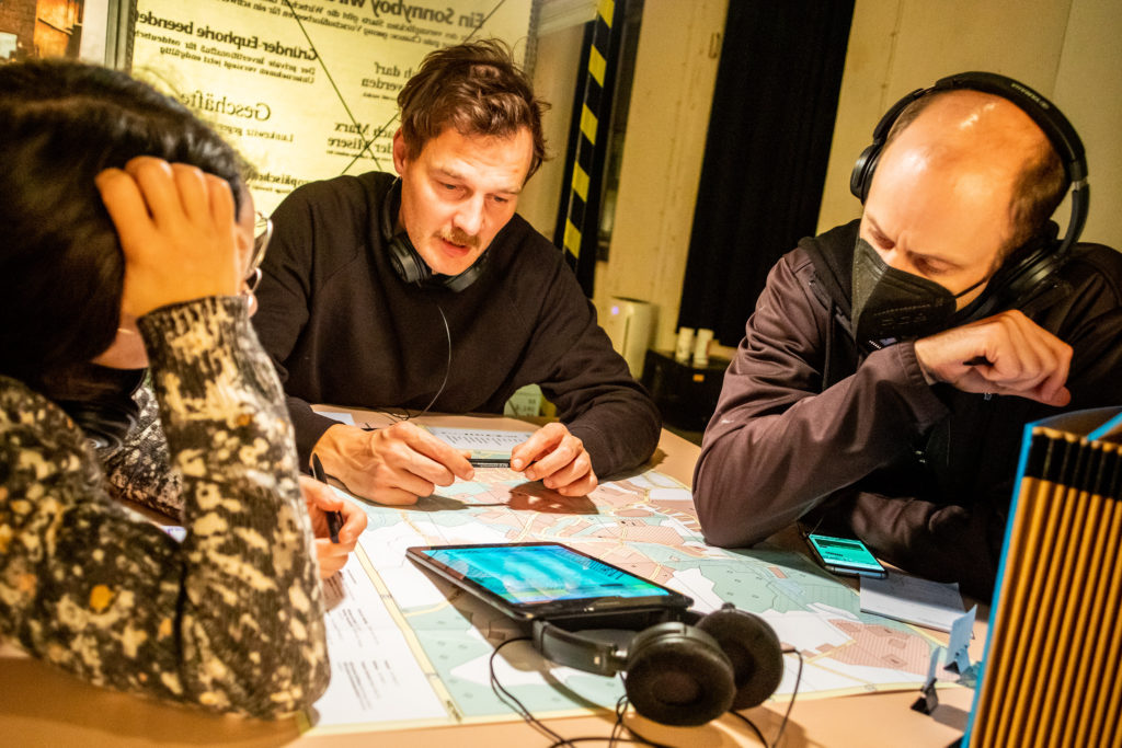 On a table are a city map, smartphones, headphones, and a tablet. Three people with headphones are sitting at the table, one person is reading something from the tablet.