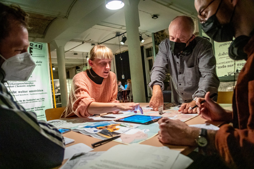 On a table are a city map, smartphones, a tablet, and printed sheets of paper. Four people sit or stand around the table, one of them uses the tablet.