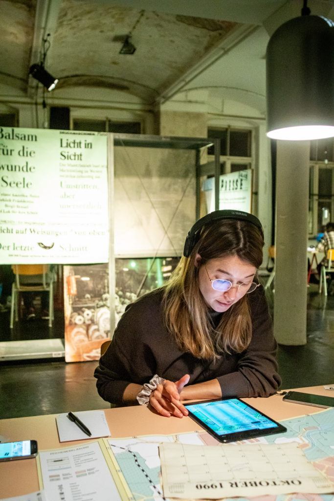 A person with long hair and glasses is sitting at a table and reading something on a tablet. In front of her are a city map, smartphones, a notepad, and printed sheets of paper.