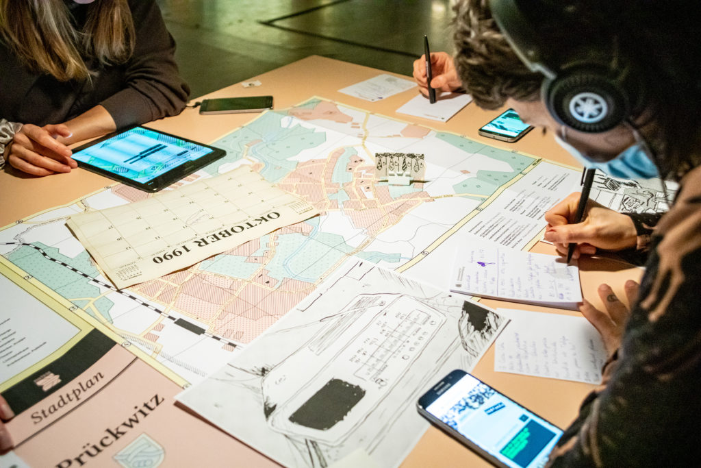 The contents of a table are visible: smartphones, a map of the city of Prückwitz, printed sheets of paper, and the hands of three people sitting around the table. Two of whom are taking notes.