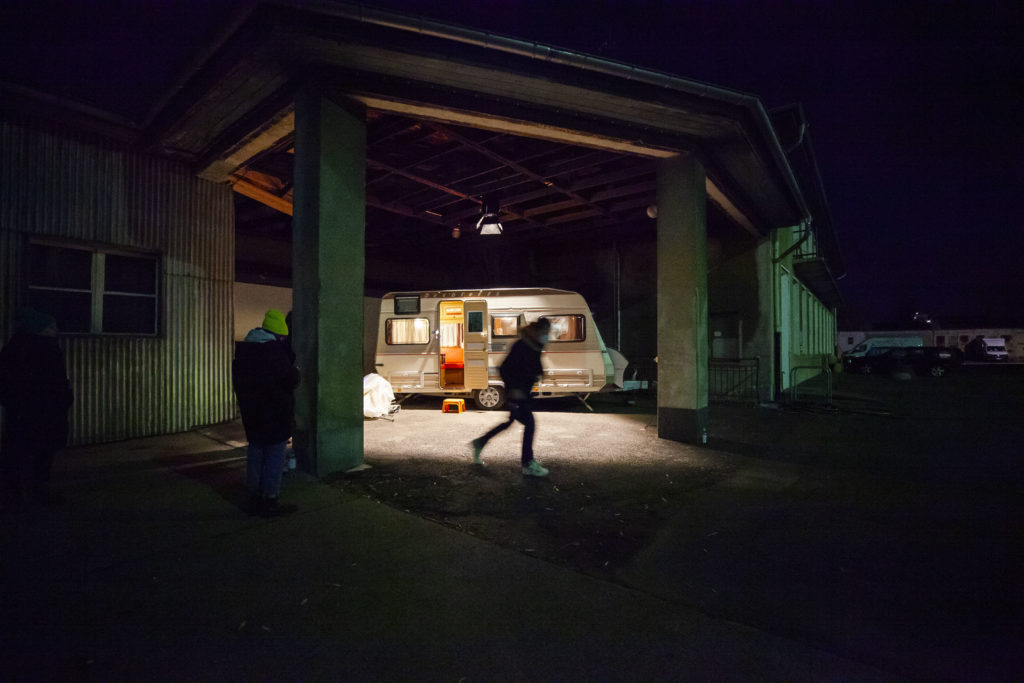 A motor home under a shelter in a dark courtyard, illuminated by a spotlight. The door is open, and the interior lit, with a silhouette in front.