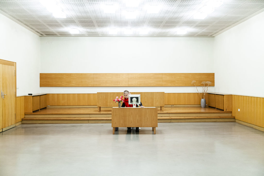 A person sits at a draped desk in the middle of a large, grand, otherwise empty room. On the desk are flowers and a screen.