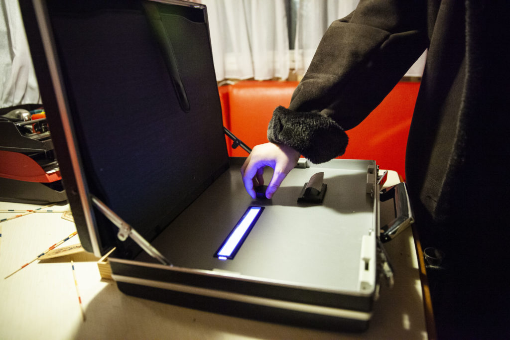 On a table is an open briefcase with a built-in receipt printer, a display, and a knob. One hand grips the knob.