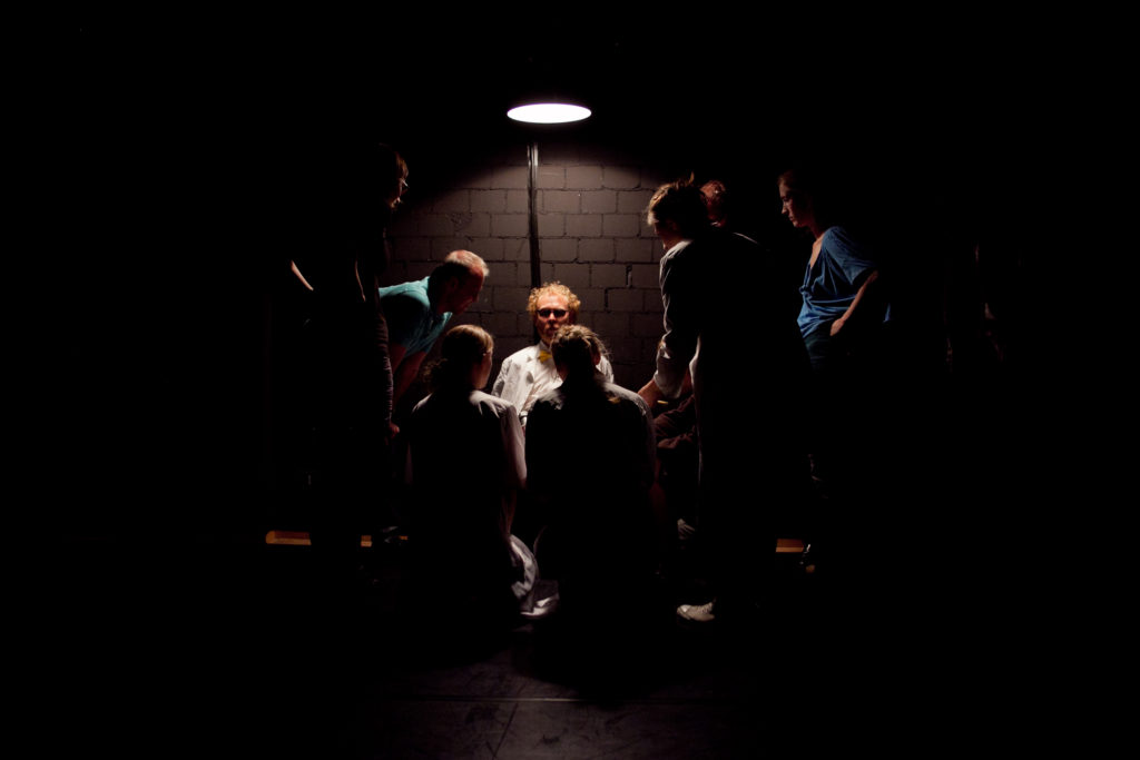 A lamp is shining on a person in a lab coat. People are standing around her in the dark, facing her.