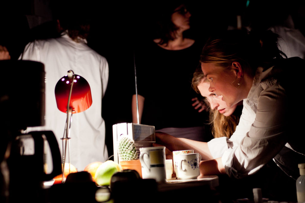 Two people bend over a table with a portable radio, a lamp, a cactus, fruit, and cups.