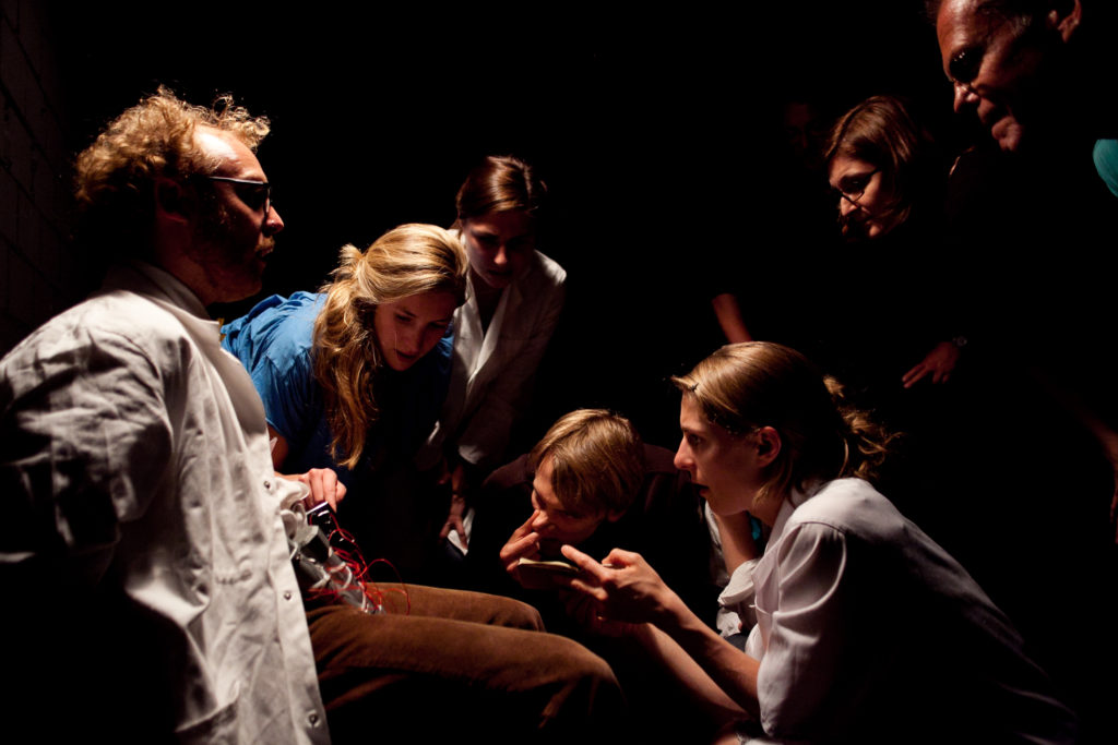 A person in a lab coat is sitting on a chair with something electronic in their lap. A group of people stands around, looking at the electronic device.