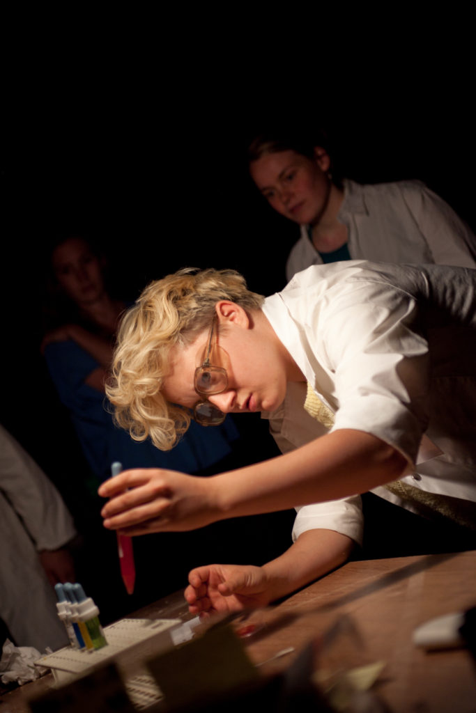 A person in a lab coat and safety goggles leans over a table and takes a test tube from a stand.