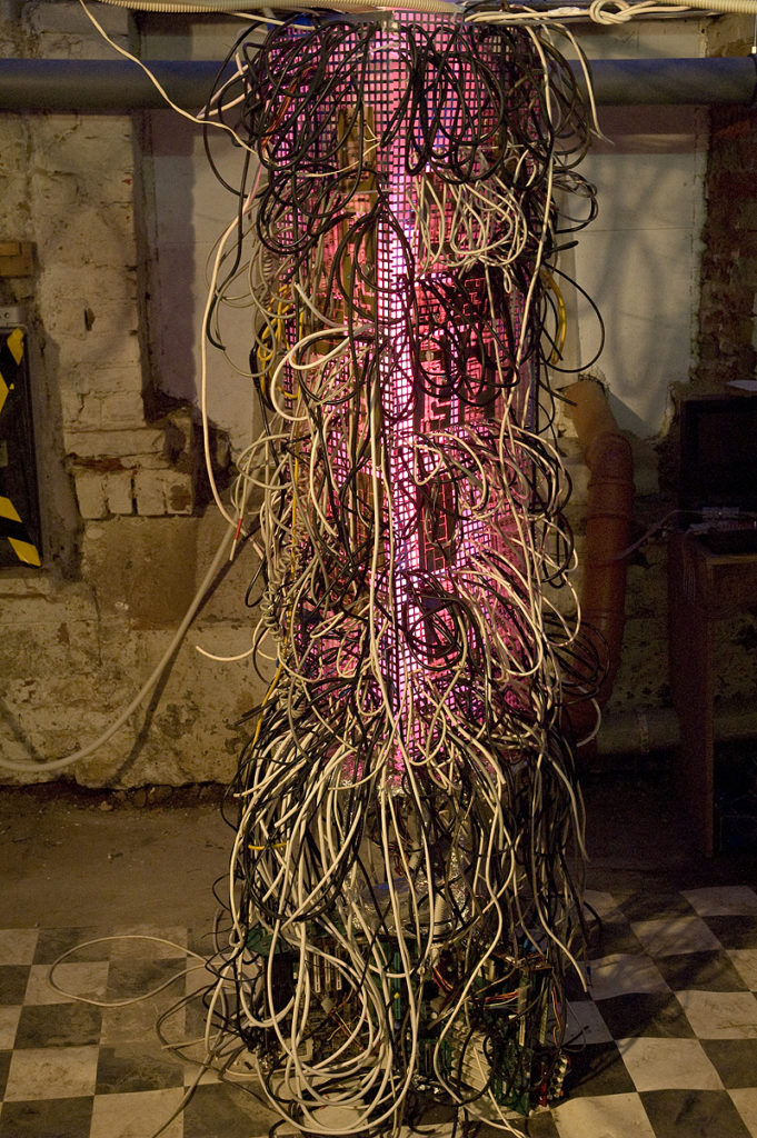 Auf schwarzweiß gekacheltem Boden steht ein pink leuchtendes Drahtgeflecht, aus dem sehr viele Kabel ragen.