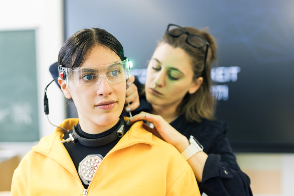 Eine weiblich gelesene Person mit einer Sicherheitsbrille wird von einer anderen weiblich gelesenen Person mit einem Schraubenzieher repariert.