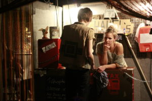 Two people stand at an improvised counter. One holds a dish towel and looks contemplative.