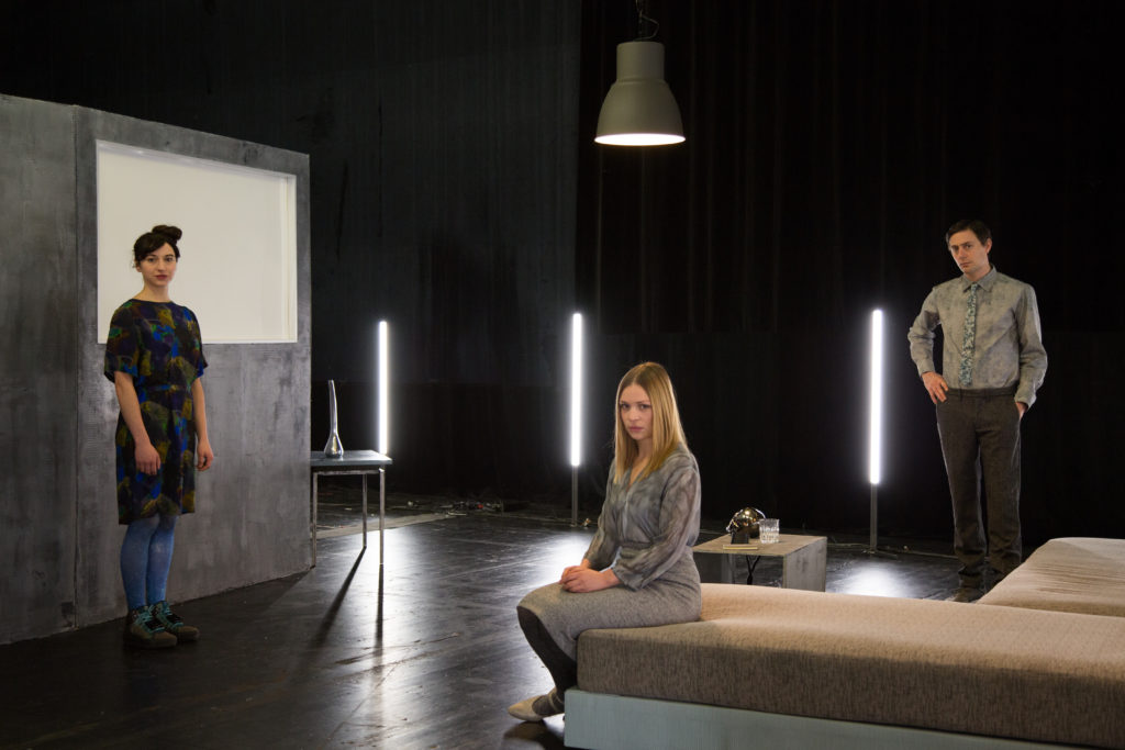 The actors Nora Decker, Ayana Goldstein, and Roland Bonjour stand in costume on a stage set and look seriously into the camera.