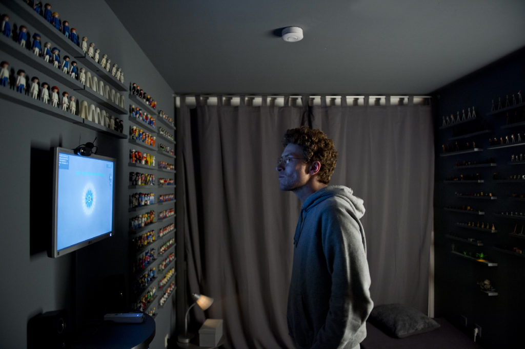 A person in a hooded sweatshirt stands in front of a gray wall containing a screen and small shelves lined with many Playmobil figurines.