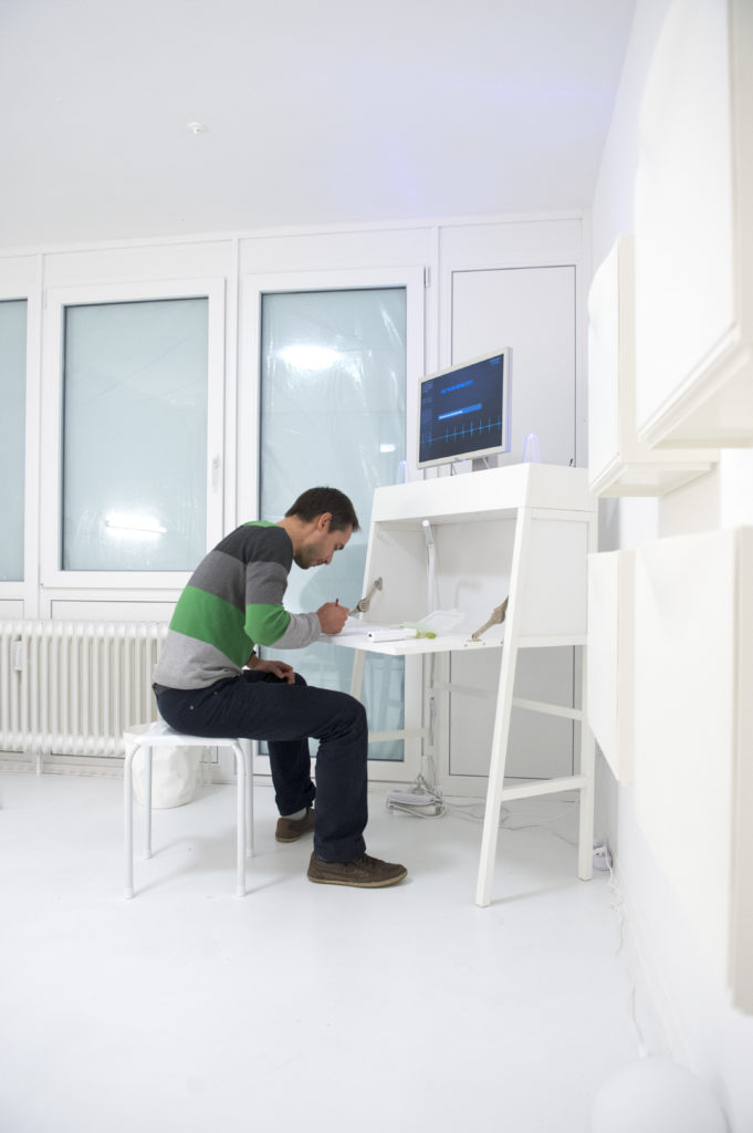 A person sits in a white room at a white desk and writes something down.