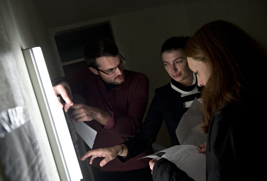 Three people hold slips of paper in their hands and look pensively at slips of paper pasted to a wall.