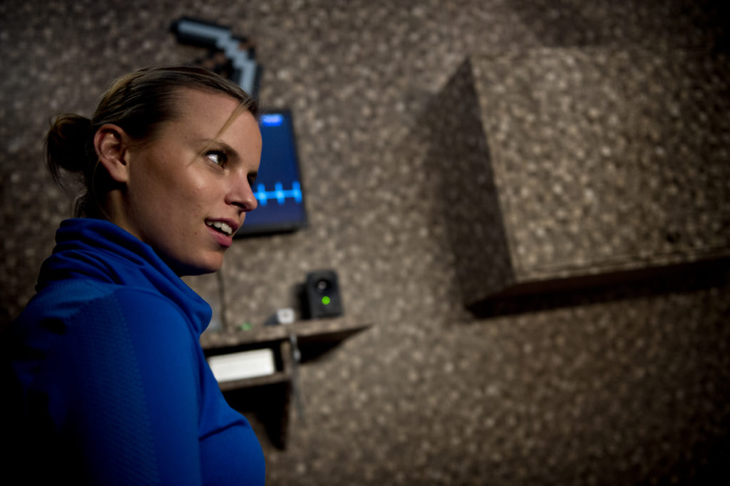 A person with their hair in a tight bun wearing a bright blue top stands in front of a wall with a screen and a loudspeaker, looking past the camera with a serious expression.