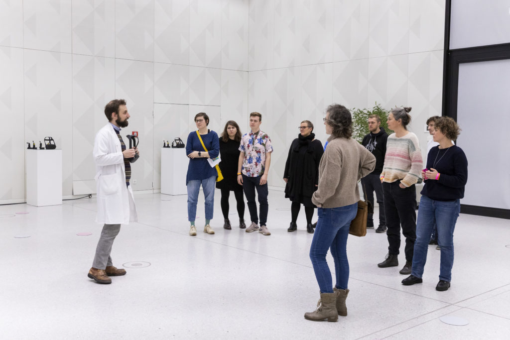 A person in a lab coat stands in front of a group of people in an all-white room and holds up a pair of VR glasses.