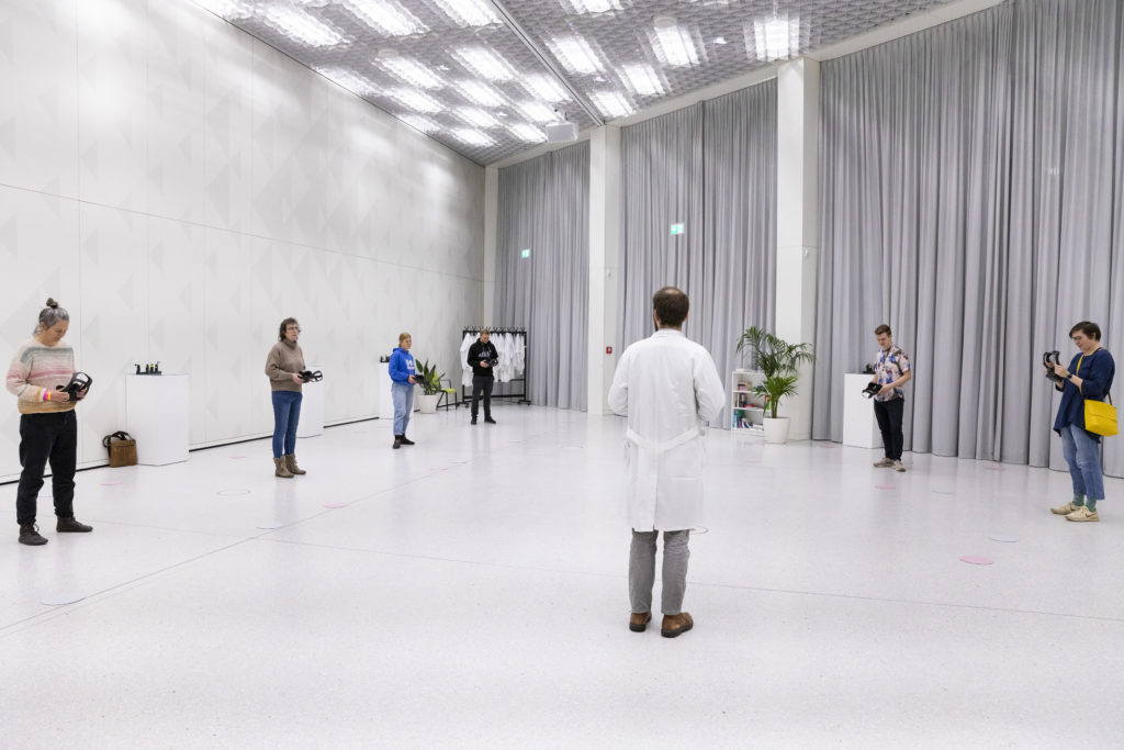 A person in a lab coat stands in an all-white room. People holding VR glasses stand around him.