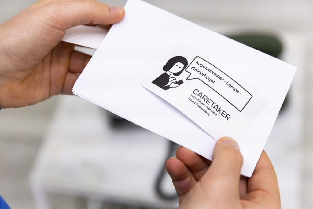 Two hands hold a business card that reads: “Pen, lamp, coat hanger. CARETAKER identification phase, Davod Rosenberg.”