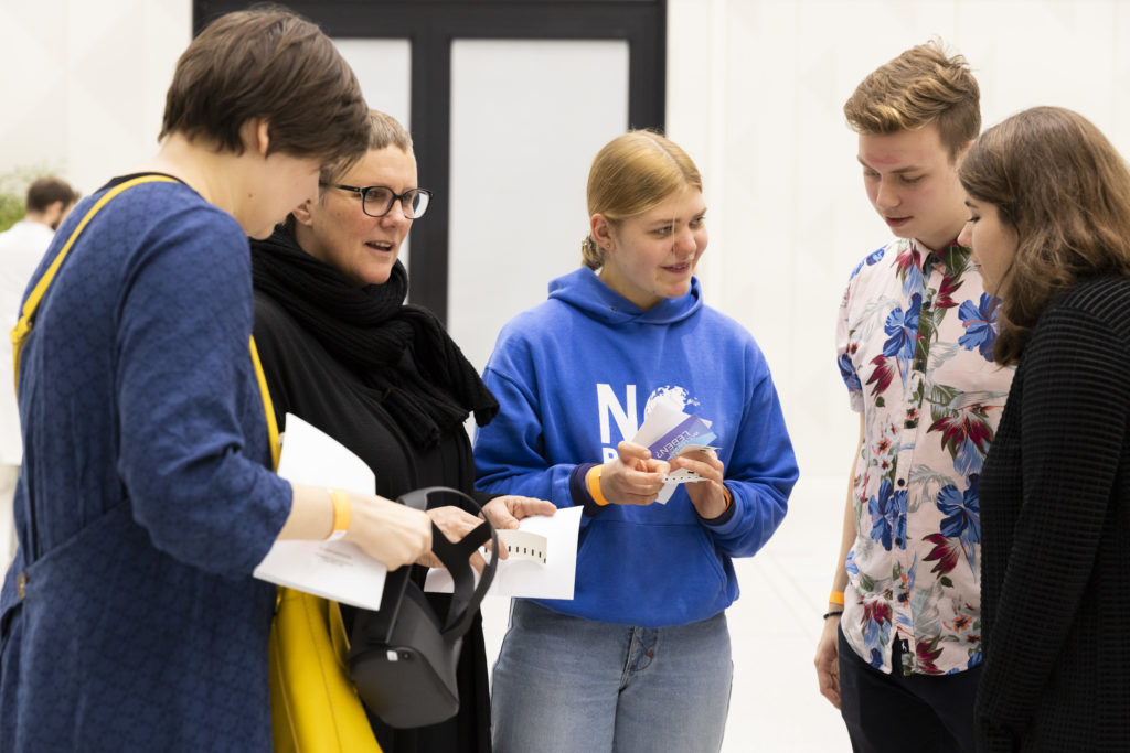 Five people are standing around together talking. They are holding tickets, paper, and VR glasses in their hands.