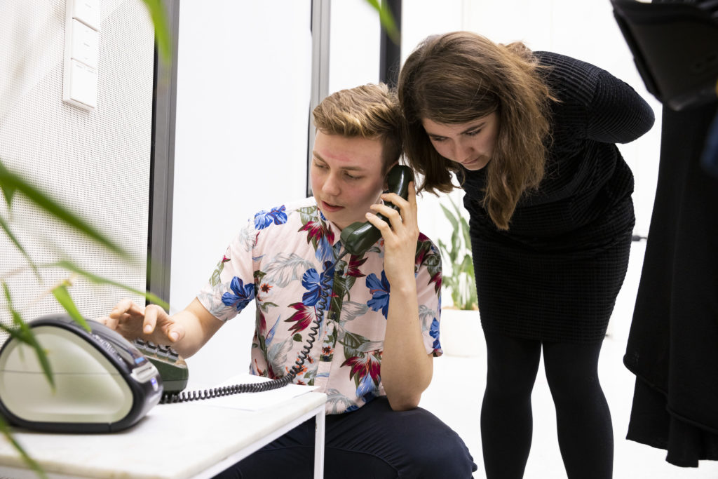 A person dials a landline telephone and holds the receiver to their ear. Another person leans over to listen in.