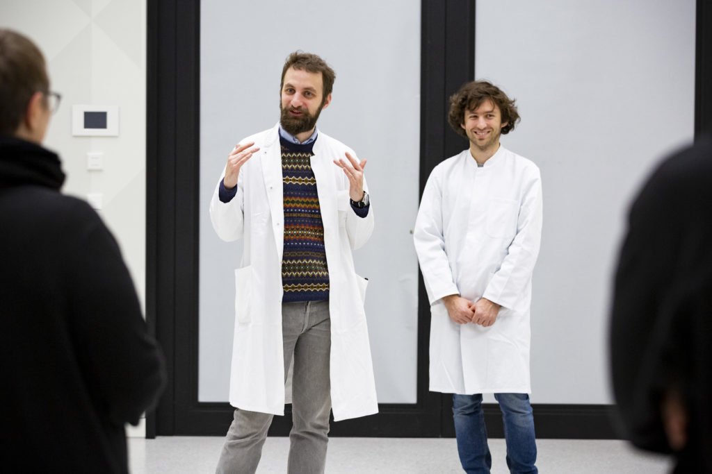 A person in a lab coat stands in front of a group of people and makes an explanatory gesture. Another person in a lab coat stands next to them and smiles.