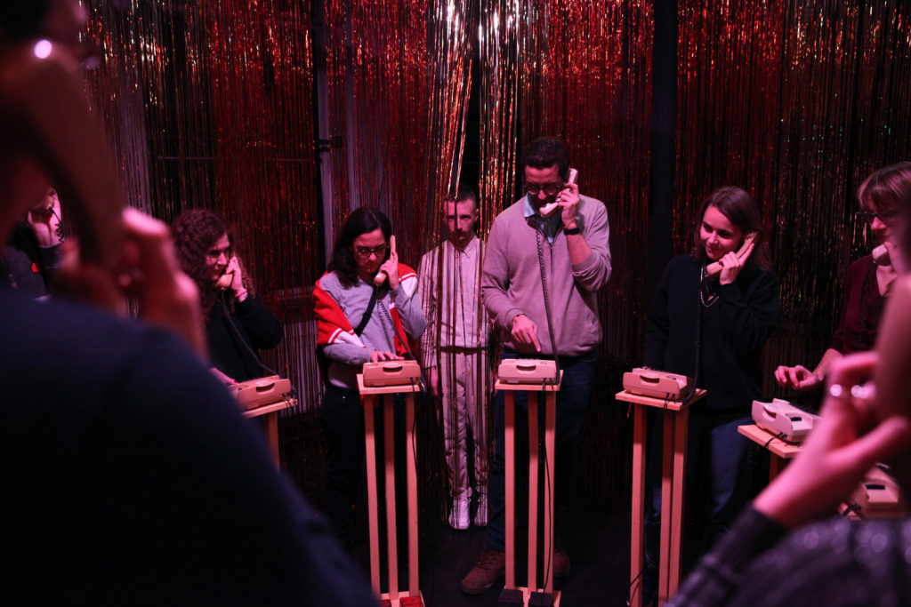 Several people stand around small standing desks with telephones, holding the phone receivers to their ears. In the background is a person behind a golden tinsel curtain.