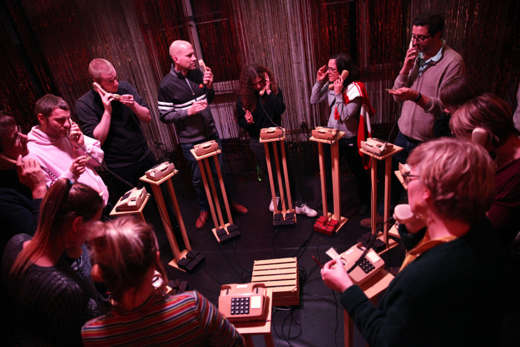 Several people stand in a circle around small standing desks with telephones, holding the phone receivers to their ears.