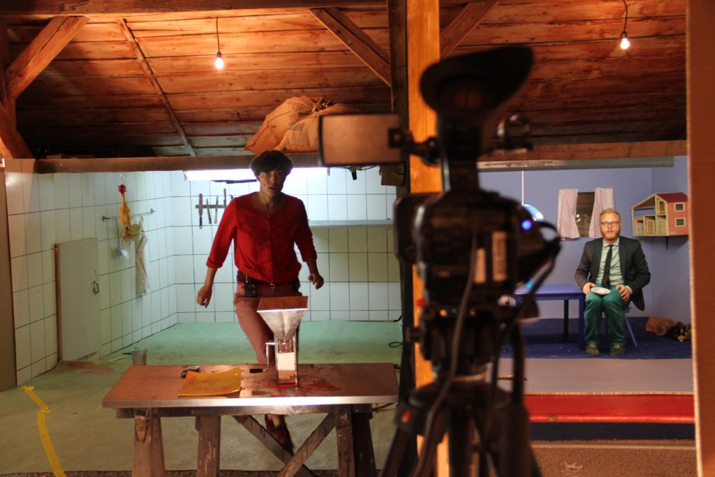 A camera in the foreground is pointed at two separate rooms in the background. In one is a person in a slaughtering room, in the other a person in a child’s room.