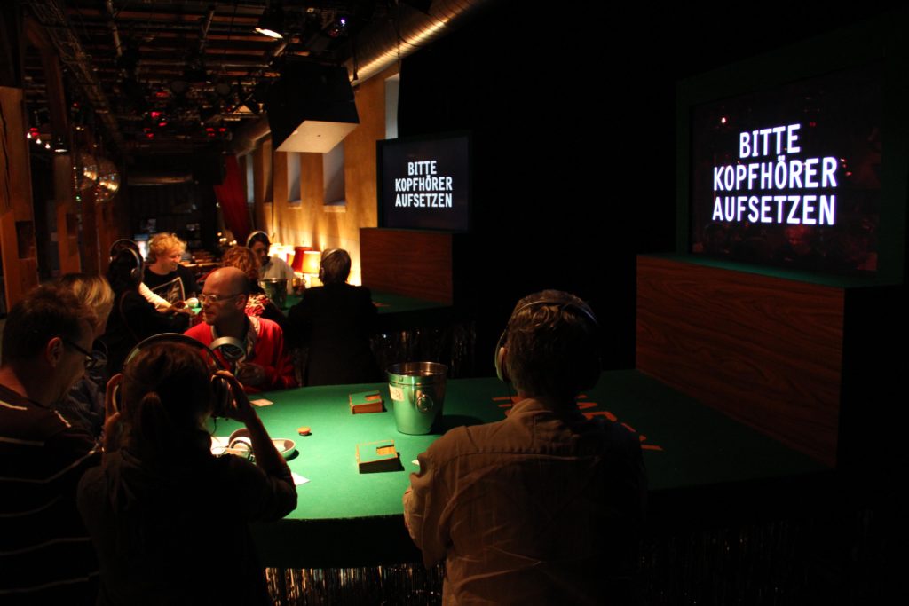 People are sitting at several casino tables. On the wall are screens that read: “Please wear headphones.”