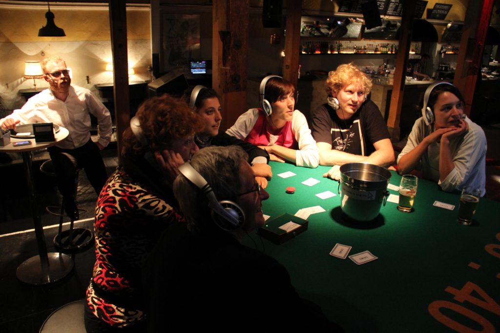 People with headphones sit at a casino table, looking transfixed at something. In the background a person in a dress shirt sits behind a glass pane.