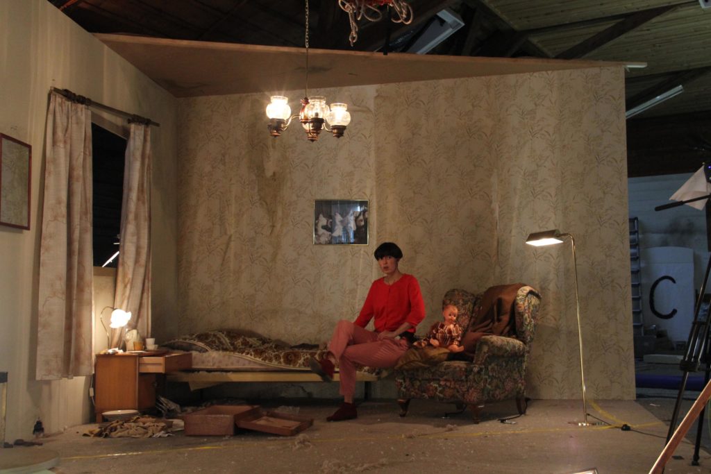 A person in red clothing sits on a bed in a retro-style room on a film set.