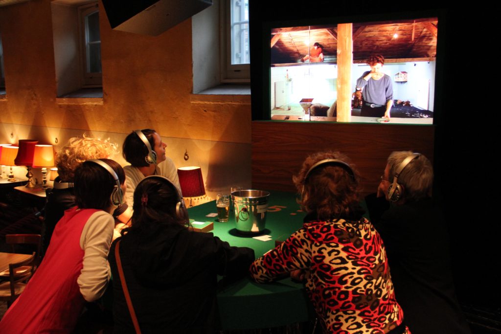 People wearing headphones at a casino table are looking at a screen showing two people in separate rooms.