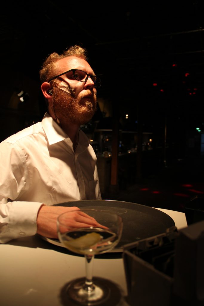 A person in a white shirt is sitting at a table. On the table are a serving tray and a glass of wine.