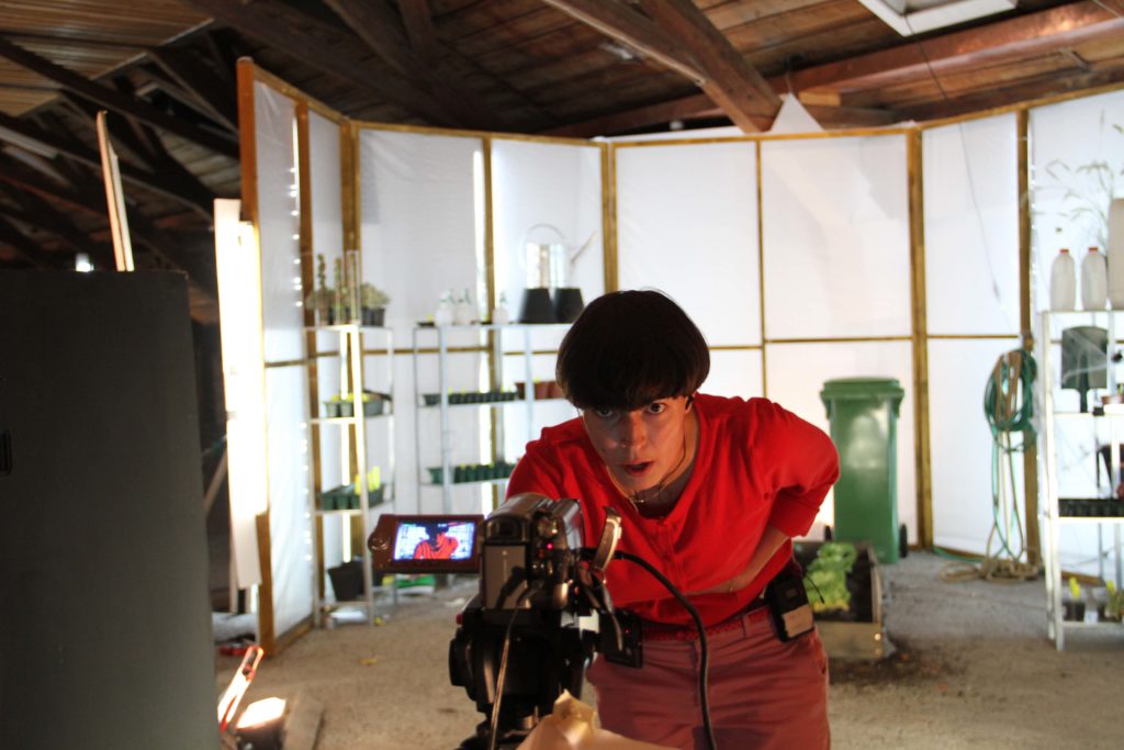 A person in a red blouse stands in front of a film set with gardening equipment and looks into a camera on a tripod.