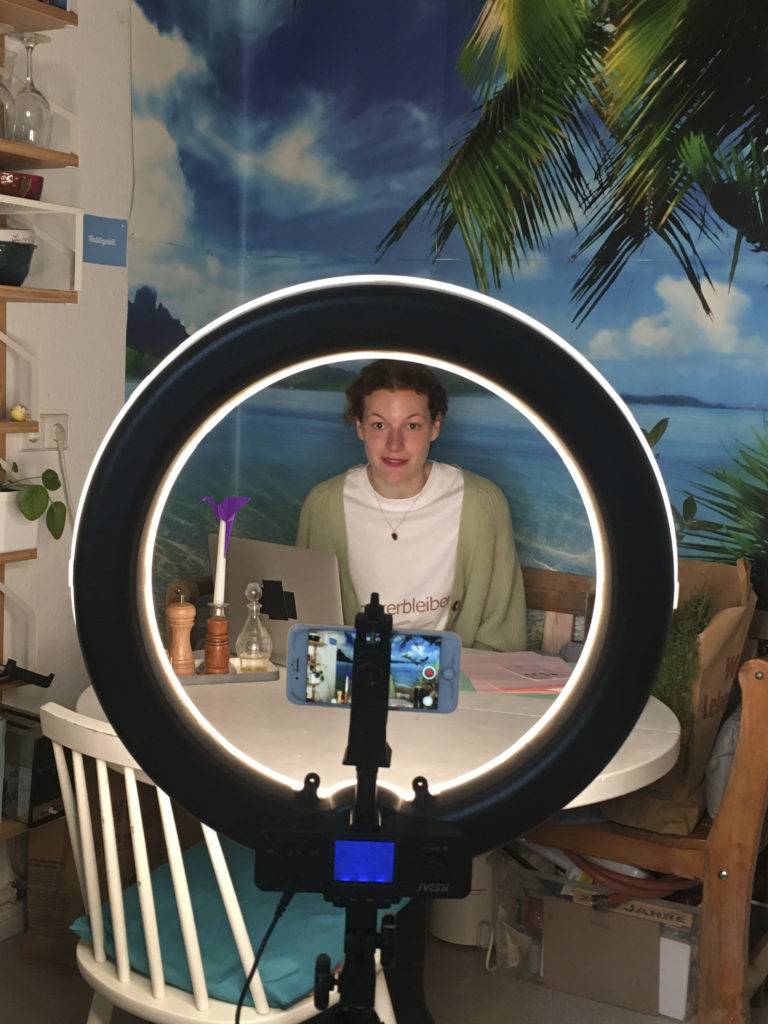 A person sits at a kitchen table in front of wallpaper with a tropical scene and looks toward a filming smartphone and a light mounted on a tripod.