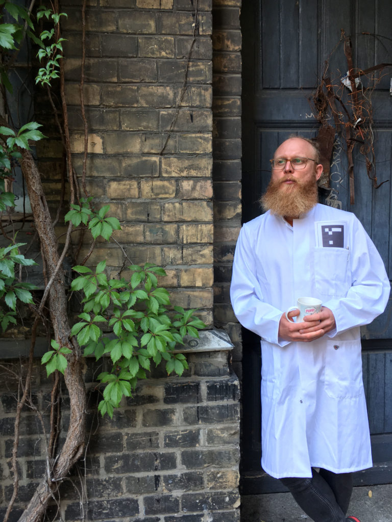 A person in a lab coat with a coffee cup in hand leans against a brick wall in front of a blue door.