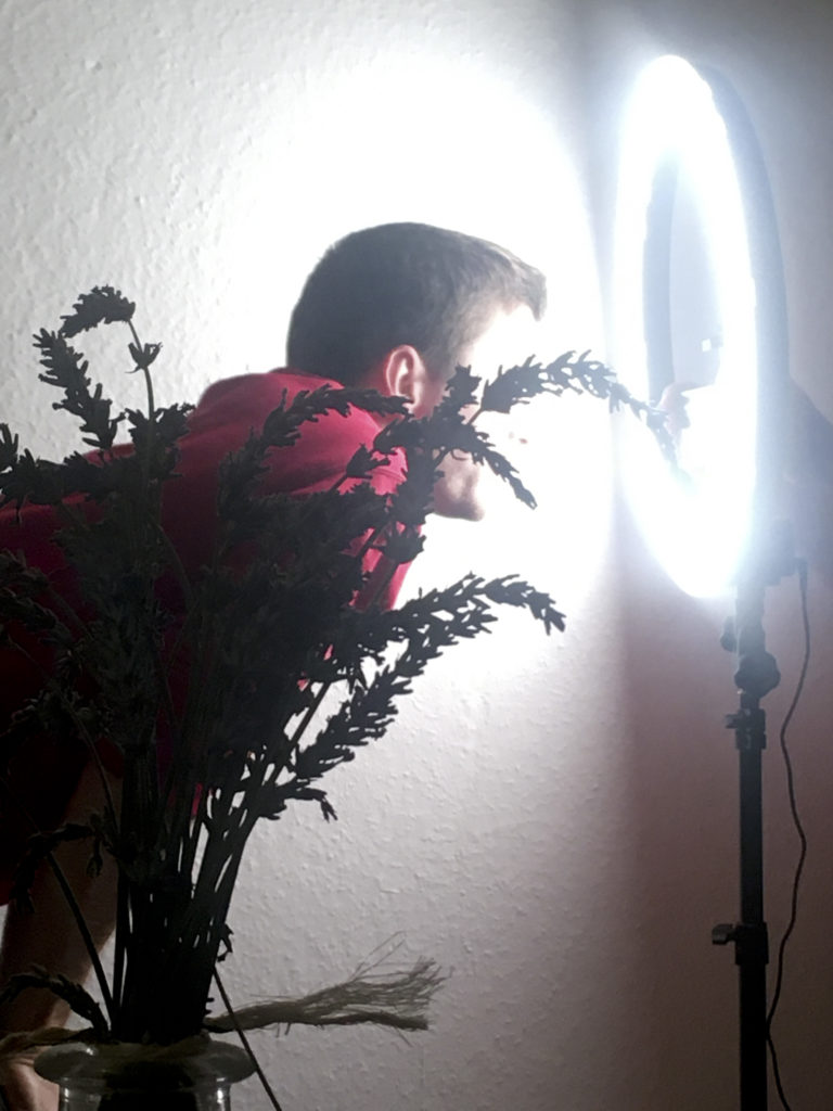 A person leans forward and stares into a circular spotlight on a tripod. She is obscured by a houseplant in the foreground.