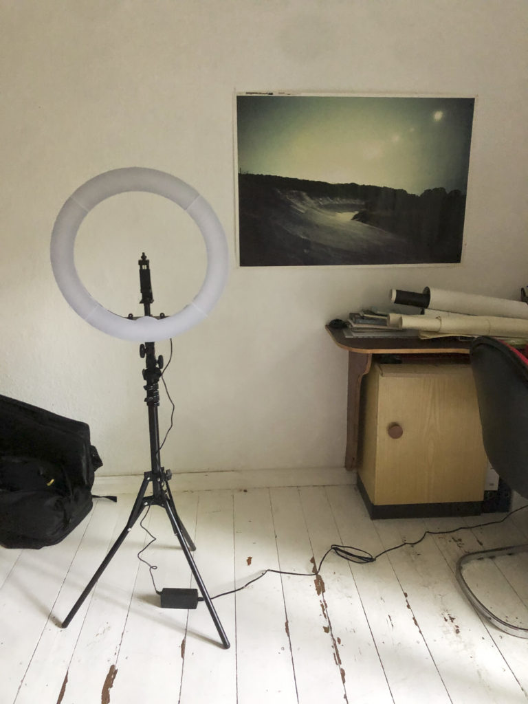 A circular light, turned off, on a tripod atop a plank. In the background a table with rolls of paper and a poster on the wall with an image of a landscape.