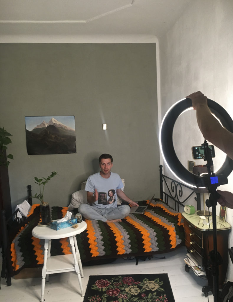 A person sits cross-legged on a bed with a bedspread and a laptop. She is illuminated by a light on a tripod that someone is operating.