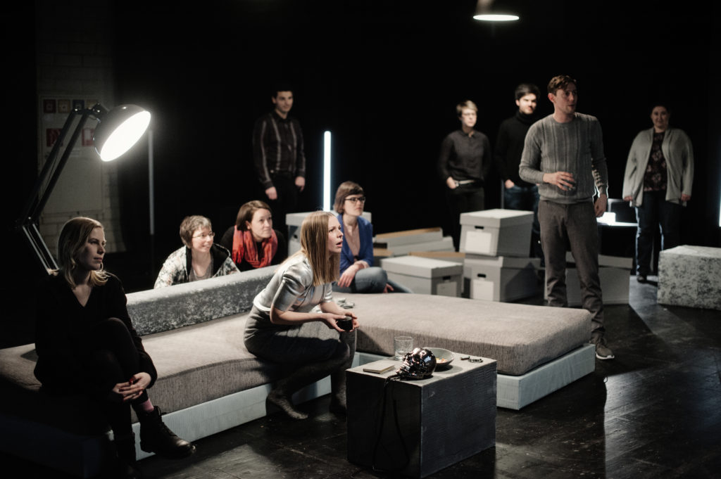 Several people are sitting on a gray sofa or standing around between gray moving boxes. All of them look in one direction, enthralled.