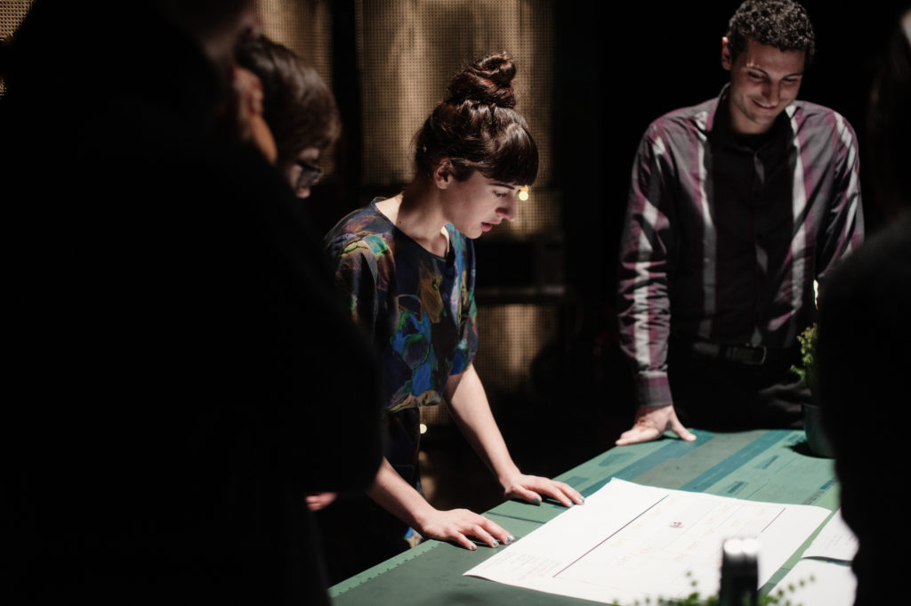 A person with bangs and her hair in a bun looks at a floorplan on the table in front of her. She is surrounded by other people.