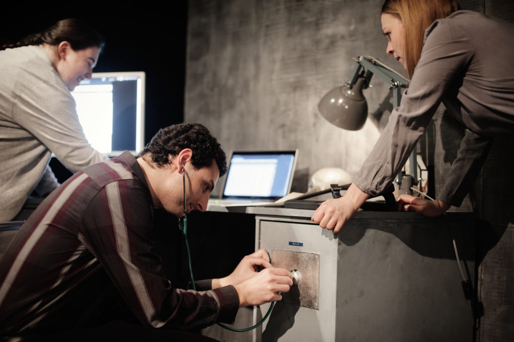 A person listens to a safe wall through a stethoscope. Next to them are two other people, laptops, and a screen.