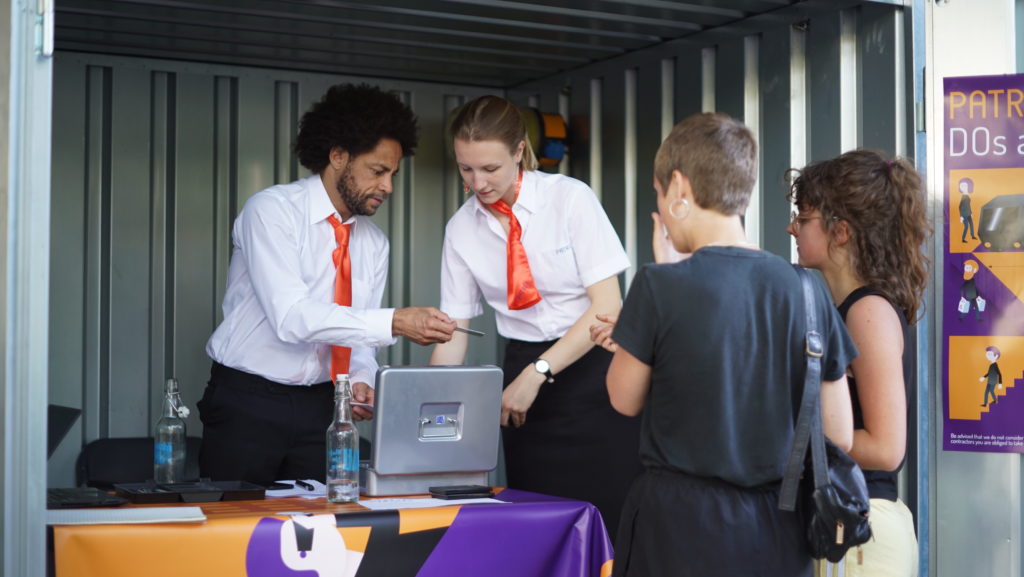 Zwei Personen in Hemden und roten Krawatten beugen sich über eine Kasse in einem offenen Baucontainer. Vor ihnen stehen zwei Personen in informeller Kleidung.