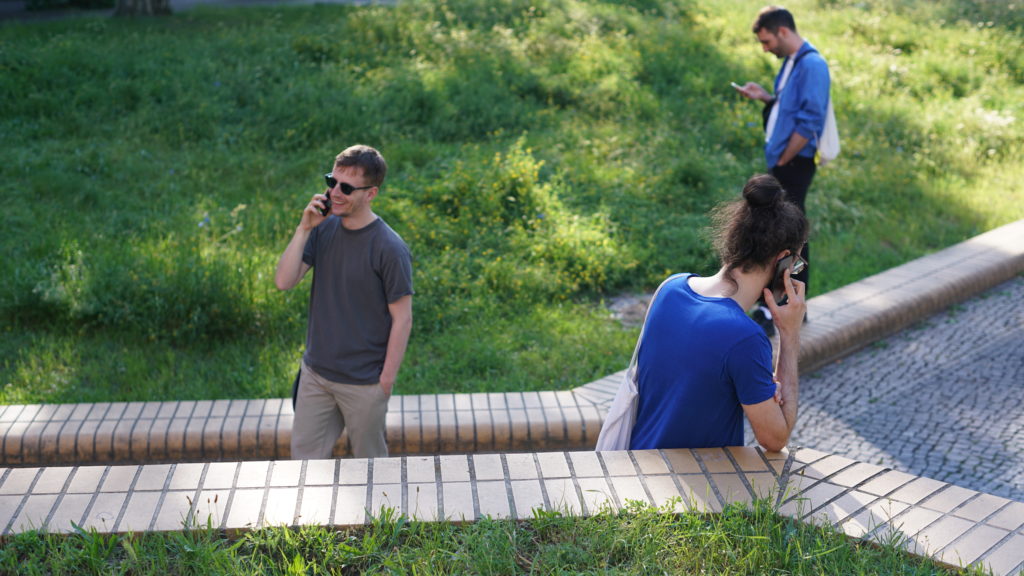 Three people are standing around in a park-like area, busy with their smartphones.