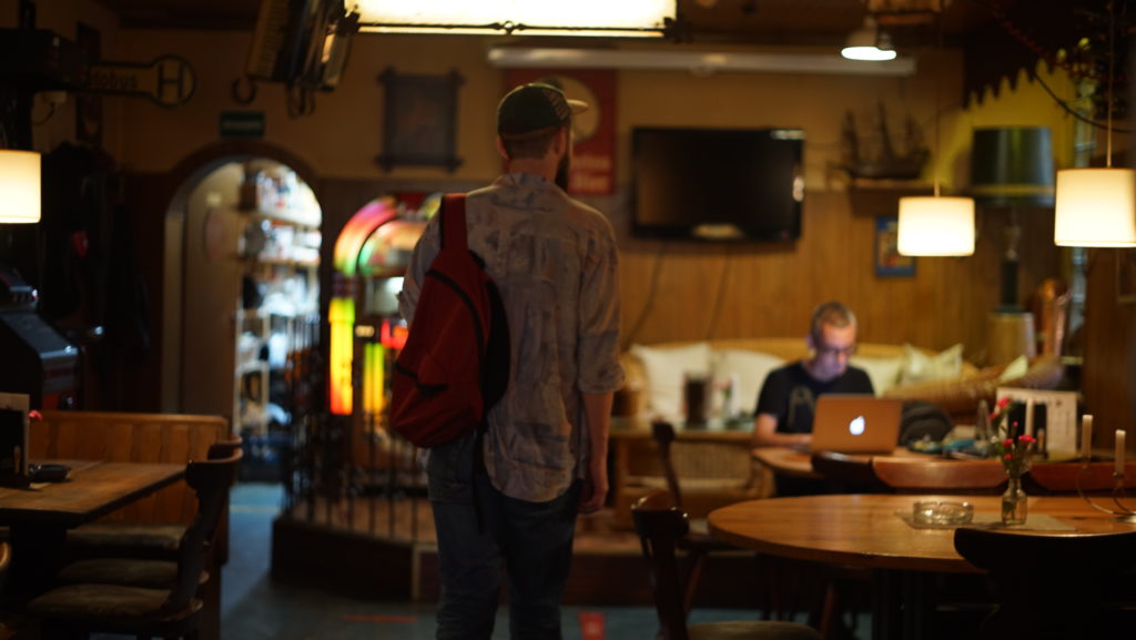 A person with a backpack approaches a person sitting at a table in a café. There is a Macbook on the table.