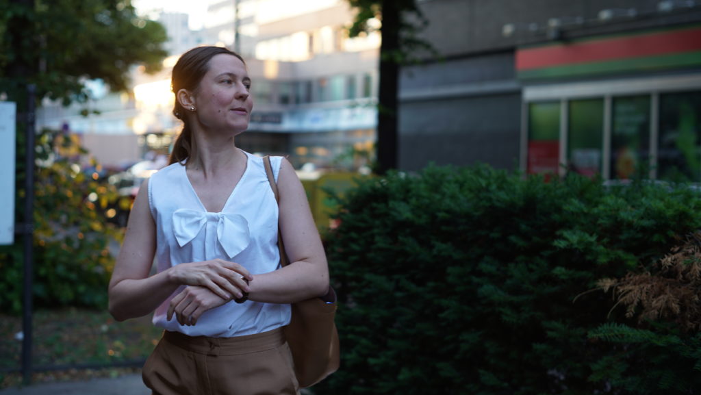 A person in a sleeveless blouse with a beige handbag stands in front of post-war urban buildings and looks out of the photo on the right.