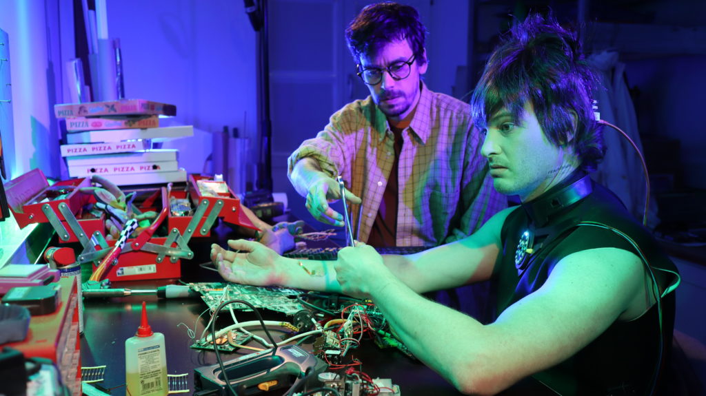 A messy desk with covered with electronics and pizza boxes. A rigid-looking person with wires in the back of their head hands a wrench to another person.