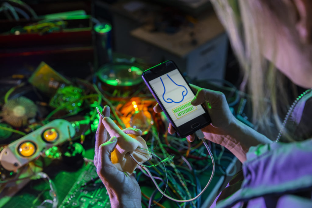 Eine Person hält eine künstlische Nase in der Hand, die mit einem Smartphone in der anderen Hand verbunden ist. Auf dem Bildschirm steht "Nasensensoren aktiviert".